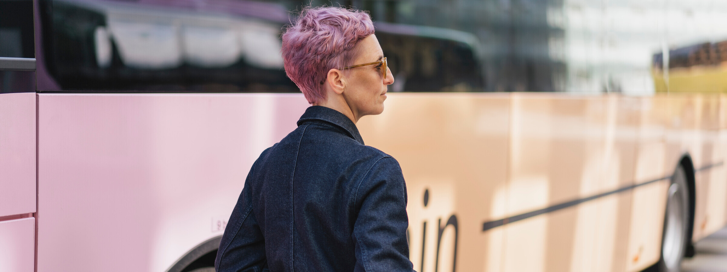 Eine Nahaufnahme einer Frau mit kurz geschnittenem, rosafarbenem Haar steht neben einem VVT Regiobus. Sie trägt eine blaue Jeansjacke und hat die Hände in den Taschen.