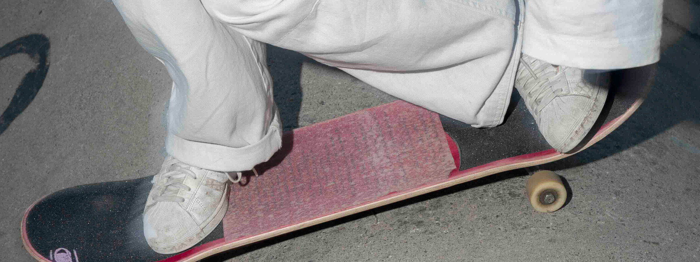 Detailaufnahme eines Skateboard-Tricks, bei dem eine Person mit weißen Turnschuhen auf dem Board steht. Die Hosenbeine sind locker über den Knöcheln aufgerollt.