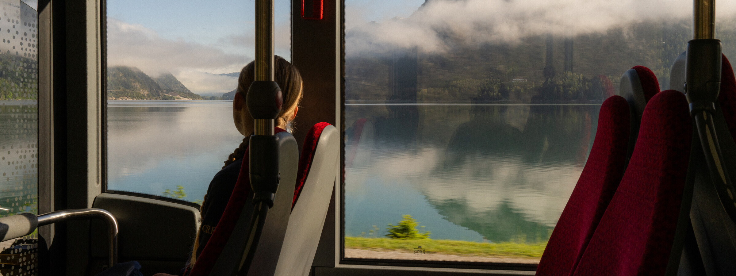 Eine Frau sitzt im Zug und schaut in eine romantische Landschaft mit See in dem sich die Berge widerspiegeln. 