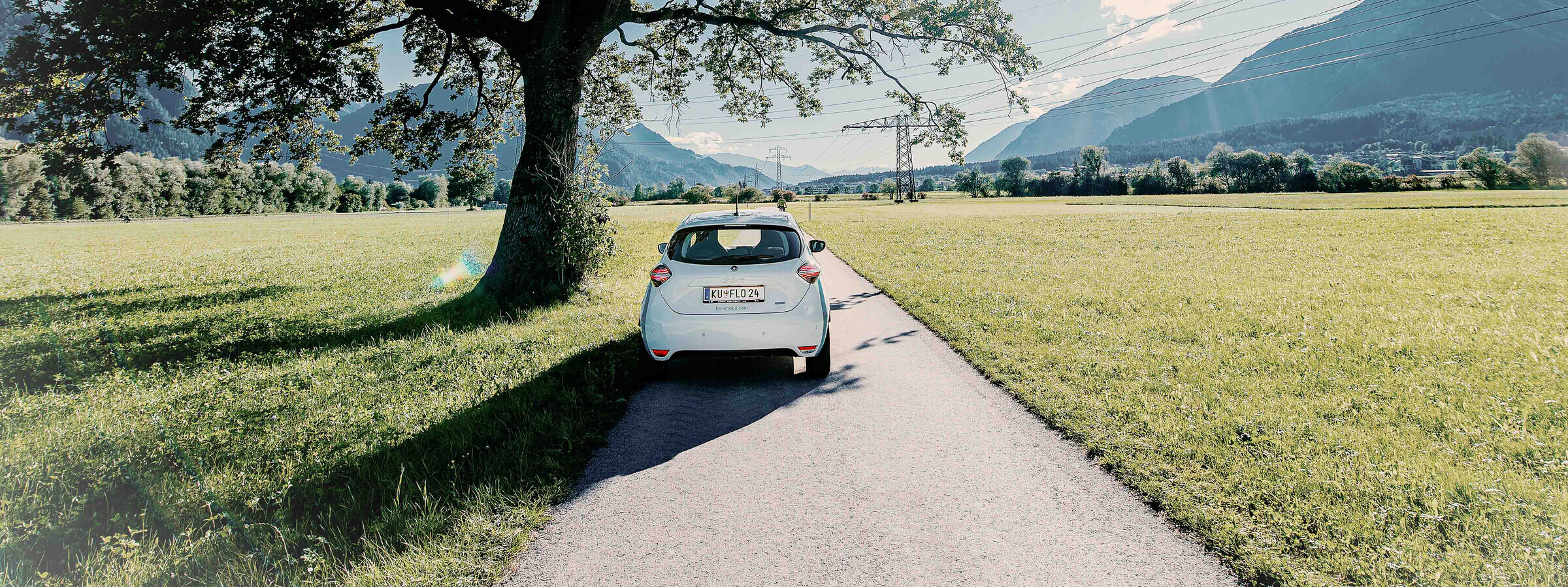 Ein weißes Auto steht auf einer schmalen, asphaltierten Straße, die durch eine grüne Wiese führt. Im Hintergrund sind Berge und Bäume zu sehen, während das Auto im Schatten eines großen Baumes parkt.