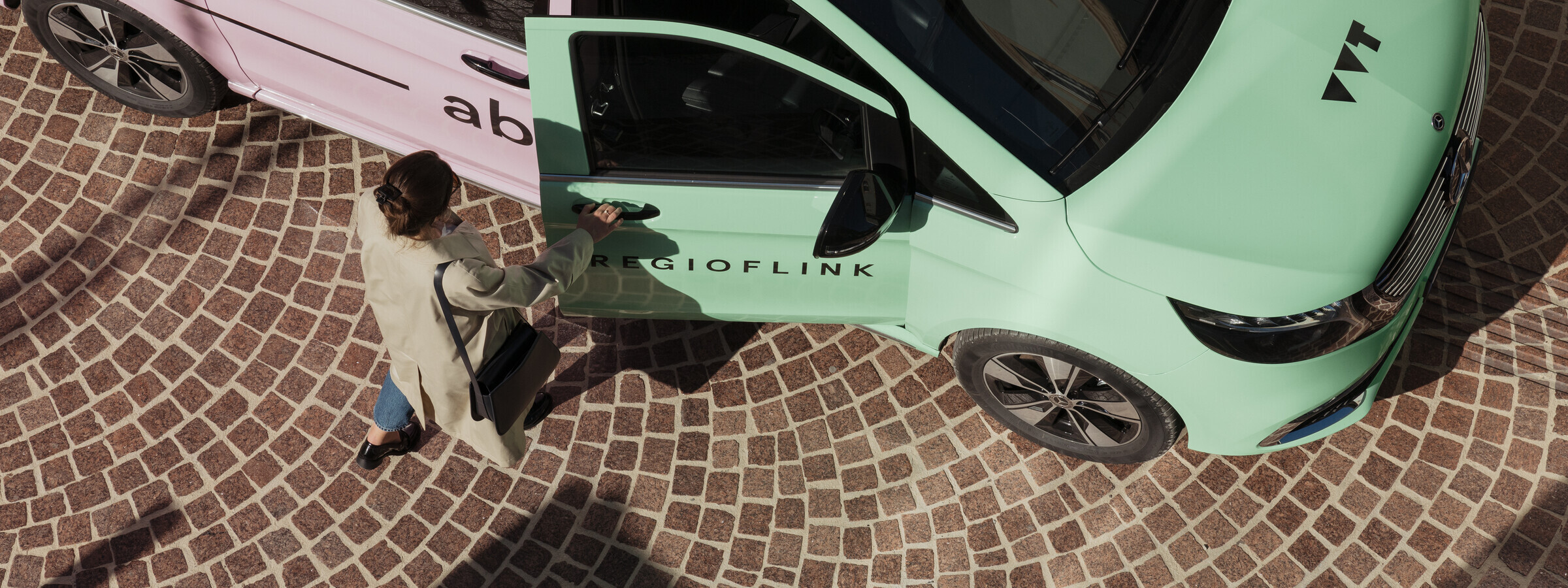 Zu sehen ist eine schicker gekleidete Frau, welche in ein bunt lackiertes RegioFlink Auto des VVT einsteigt. Das Foto ist von oben aufgenommen und das Auto steht auf gepflasterter Straße. 