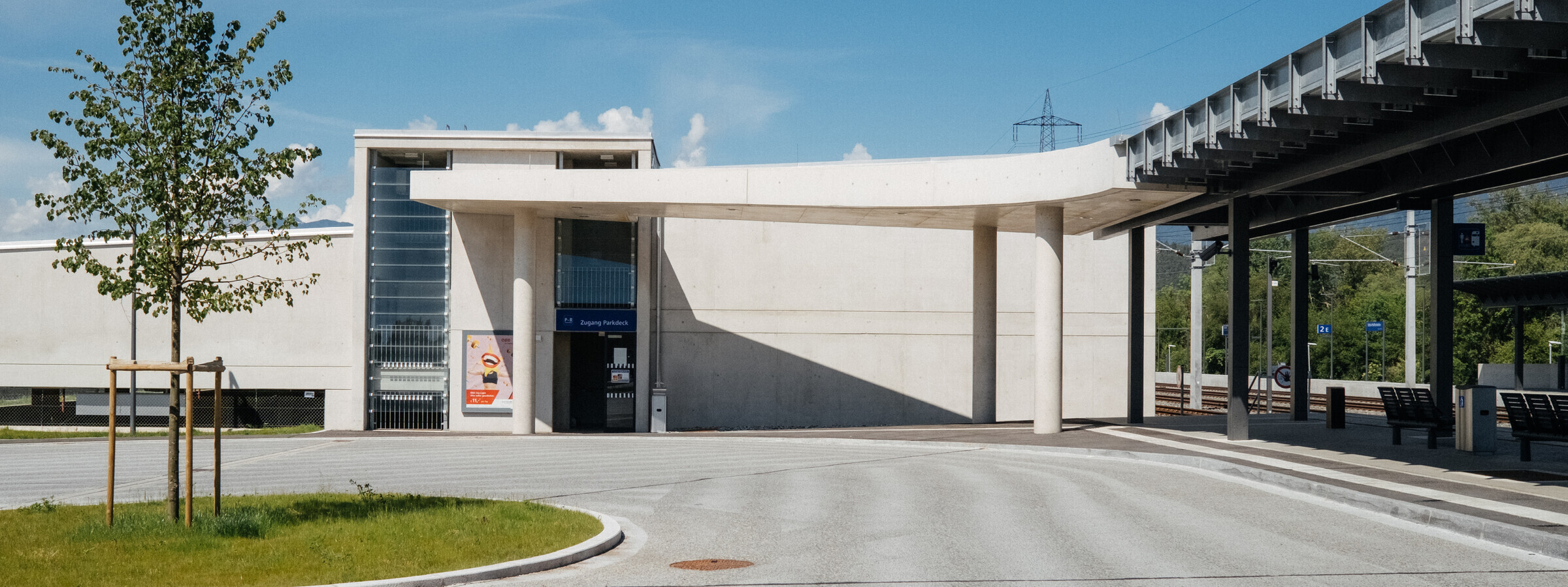 Eine Bahnstation mit einem modernen weißen Gebäude und einer überdachten Passage. Der Bereich ist sonnig und ruhig, mit einem neu gepflanzten Baum an der Seite.
