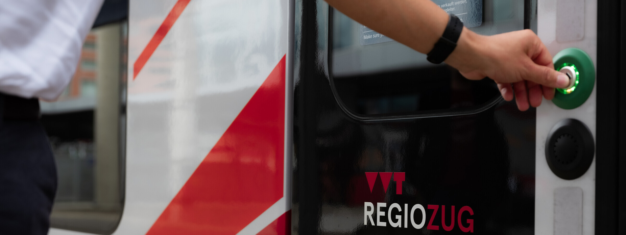 Detailansicht einer Hand, die den grünen Knopf eines VVT RegioZuges drückt, um die Tür zu öffnen. Auf der Tür ist das VVT-Logo und der Schriftzug "RegioZug" zu sehen.