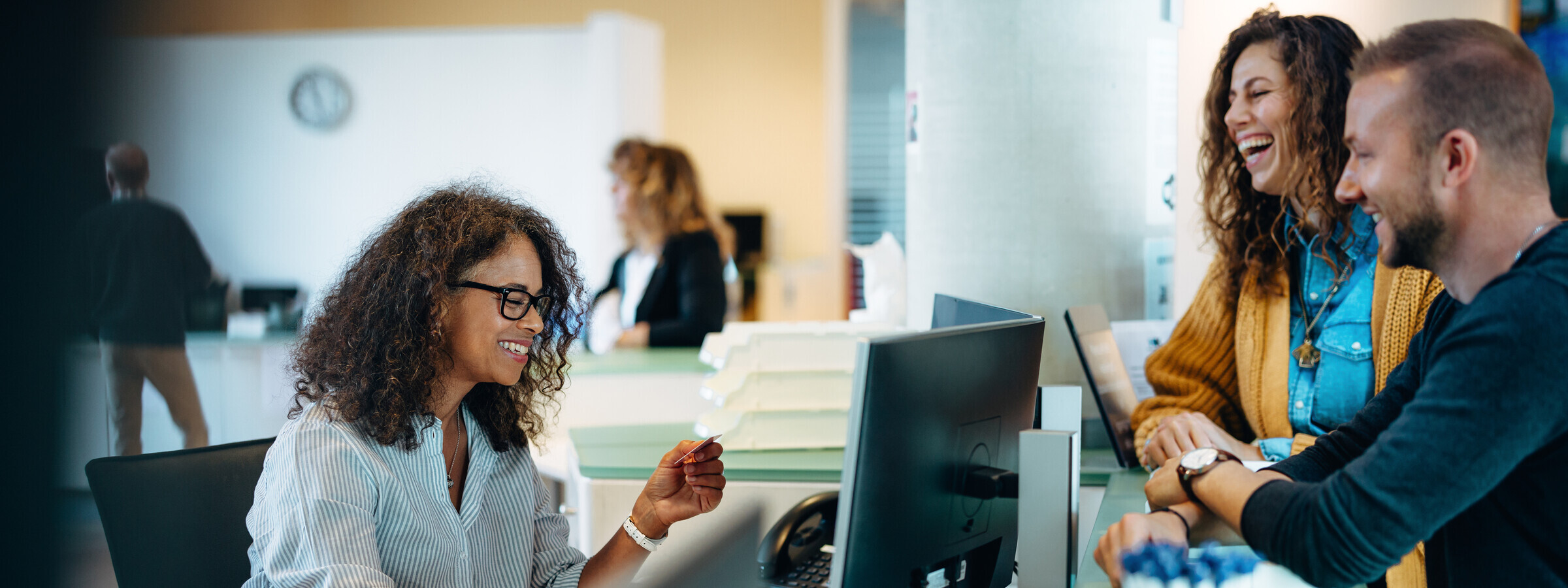 Eine Frau mit lockigem Haar und Brille sitzt lächelnd an einem Schreibtisch in einem Kundenzentrum und berät ein junges Paar, das ebenfalls lächelt. Sie befinden sich in einem modernen Empfangsbereich.