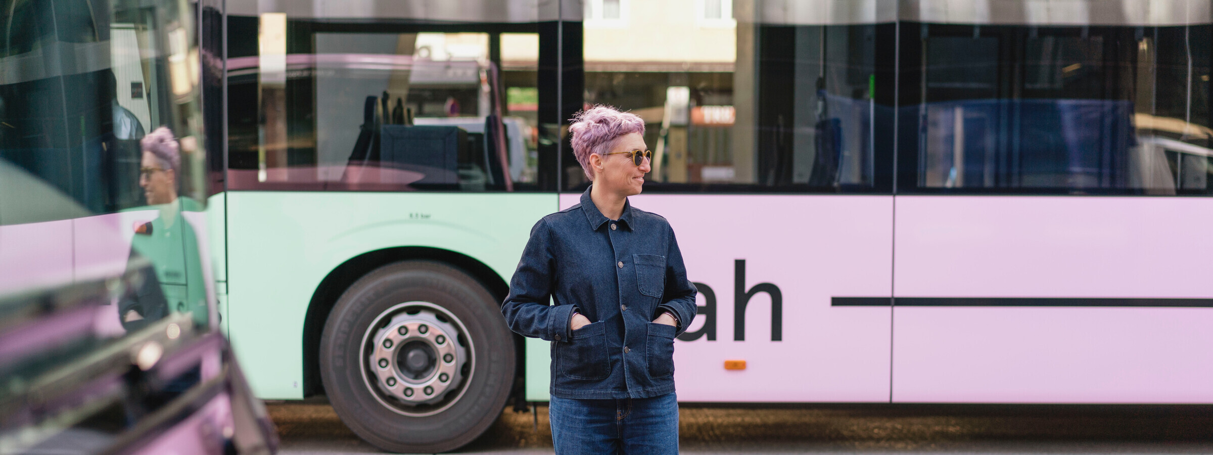 Eine Frau mit kurzem, lila Haar steht entspannt mit den Händen in den Taschen vor einem VVT RegioBus. Ihr Spiegelbild ist im Fenster eines vorbeifahrenden Busses zu sehen.