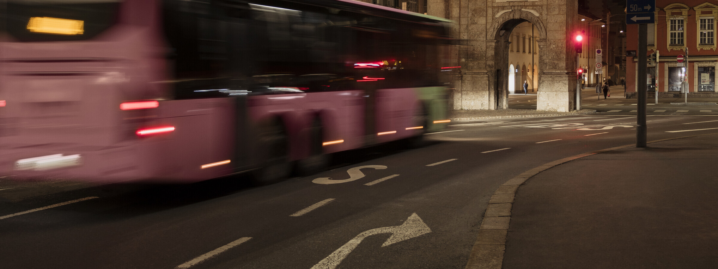 Ein unscharfer rosa VVT Bus fährt nachts durch eine leere Straßenkreuzung in der Stadt. Im Hintergrund ist ein beleuchtetes historisches Gebäude und die Triumphpforte in Innsbruck zu sehen.