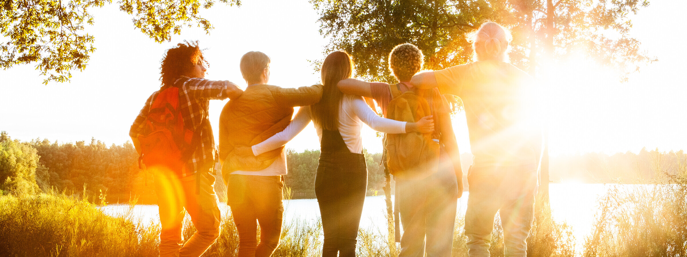 Rückansicht von fünf jungen Menschen, die sich umarmen und auf einen See bei Sonnenuntergang schauen. Das warme Licht der Sonne beleuchtet die Szene und verleiht ihr eine freundliche und einladende Stimmung.