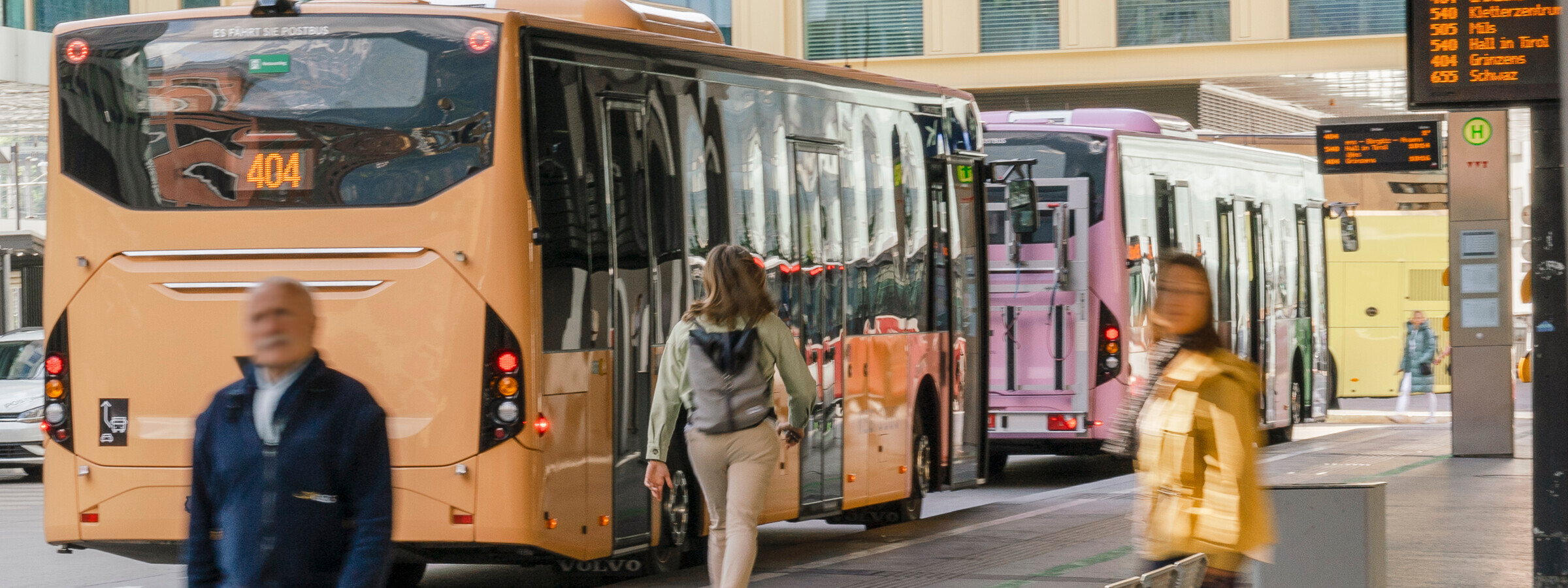 Verschiedene VVT-Busse stehen an einer Haltestelle, während Passagiere ein- und aussteigen. Die digitale Anzeigetafel im Hintergrund zeigt die Buslinien und Ziele der VVT-Busse. Im Vordergrund sind Menschen in Bewegung zu sehen.