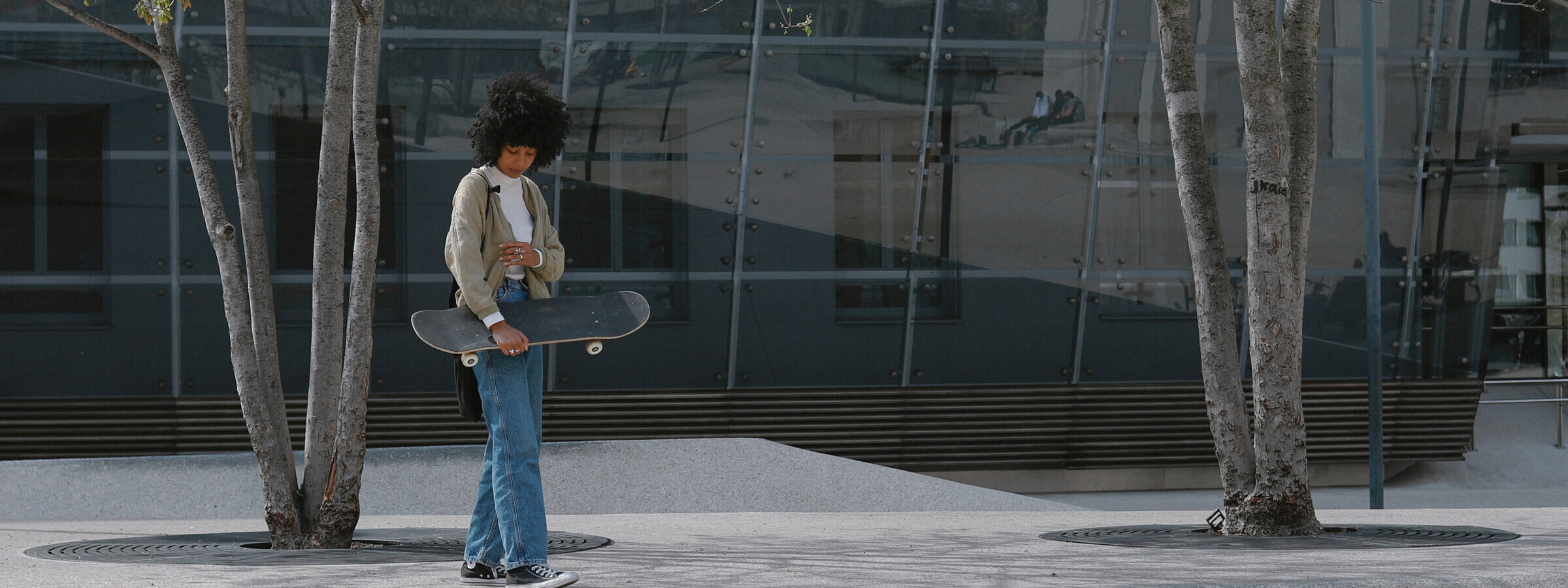 Eine Jugendliche steht mit einem Skateboard in den Händen in einem modernen städtischen Umfeld. Die Umgebung ist von einer Glasfassade und Bäumen geprägt, während die Person in lässiger Kleidung ruhig nach unten blickt.