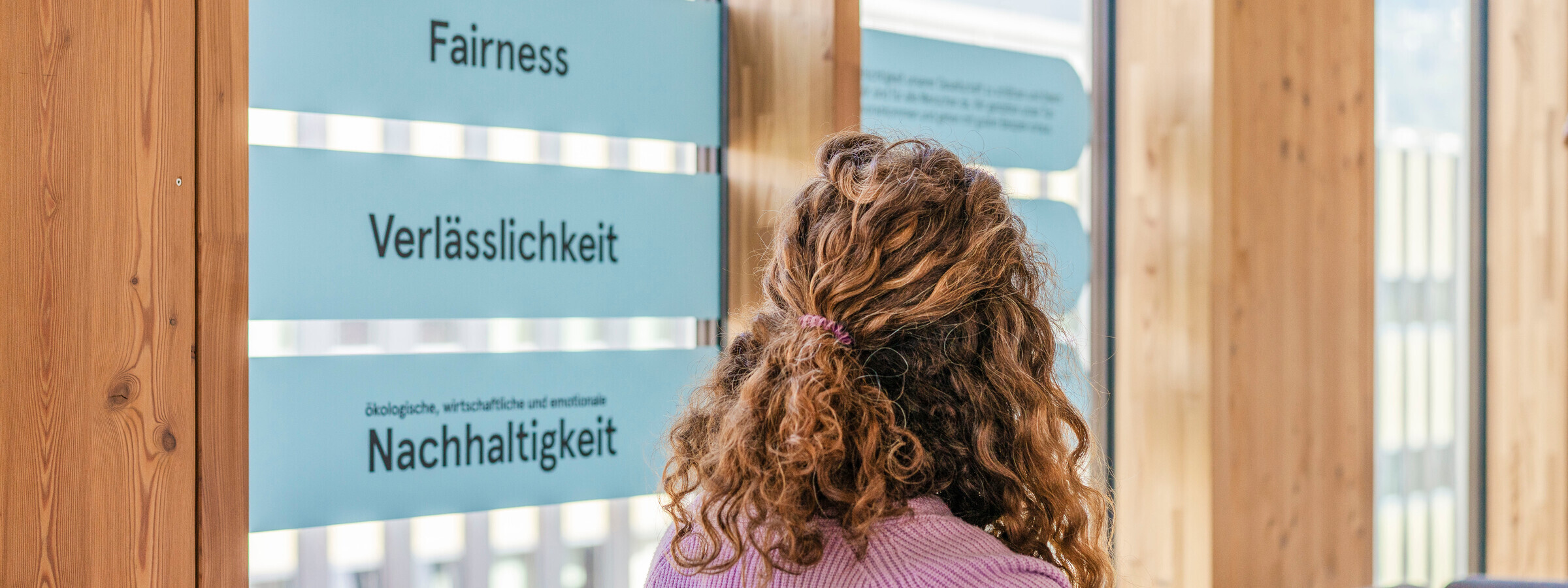 Eine Frau mit lockigem Haar blickt im VVT-Büro auf eine Wand mit den Werten "Fairness", "Verlässlichkeit" und "Nachhaltigkeit", die auf einer Glasfläche dargestellt sind. Die Holzwände und großen Fenster des Büros betonen ein nachhaltiges Design.