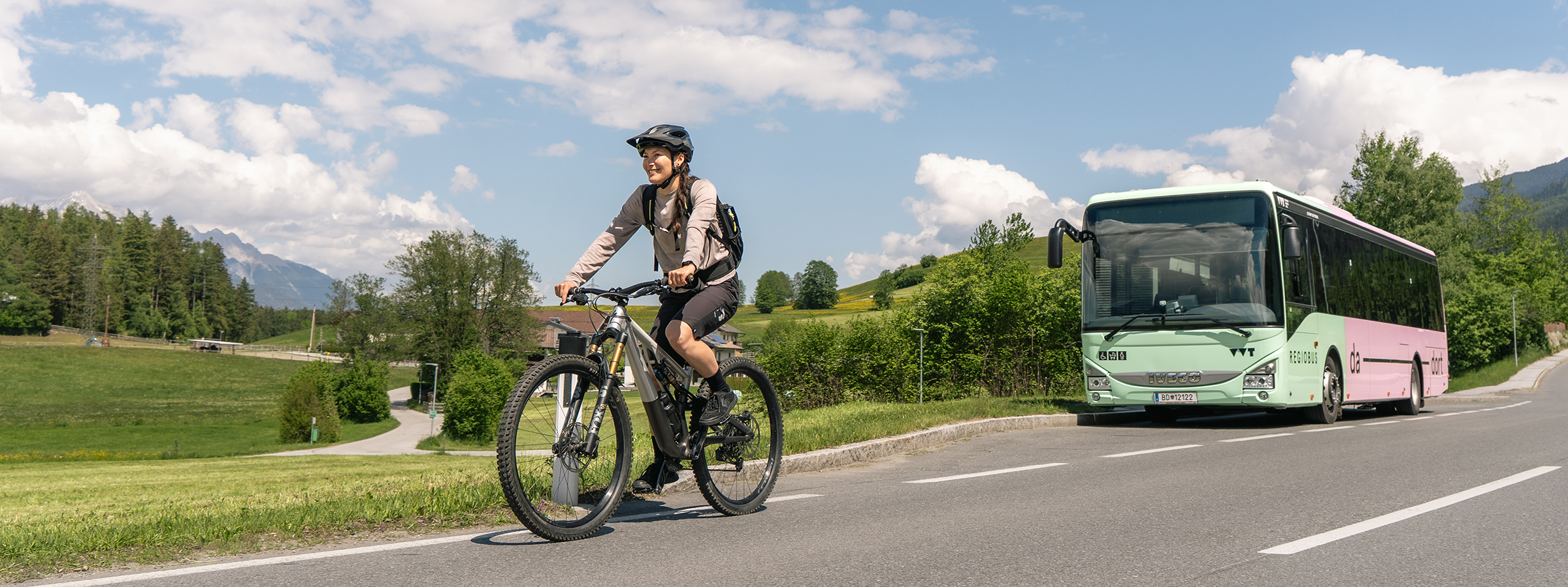 Auf dem Foto ist eine Frau in Mountainbike radelklamotten auf einem Mountainbike zu sehen, wie sie glücklich auf der Straße fährt. Im Hintergrund sieht man ein VVT Bus in den Farben grün-rosa.