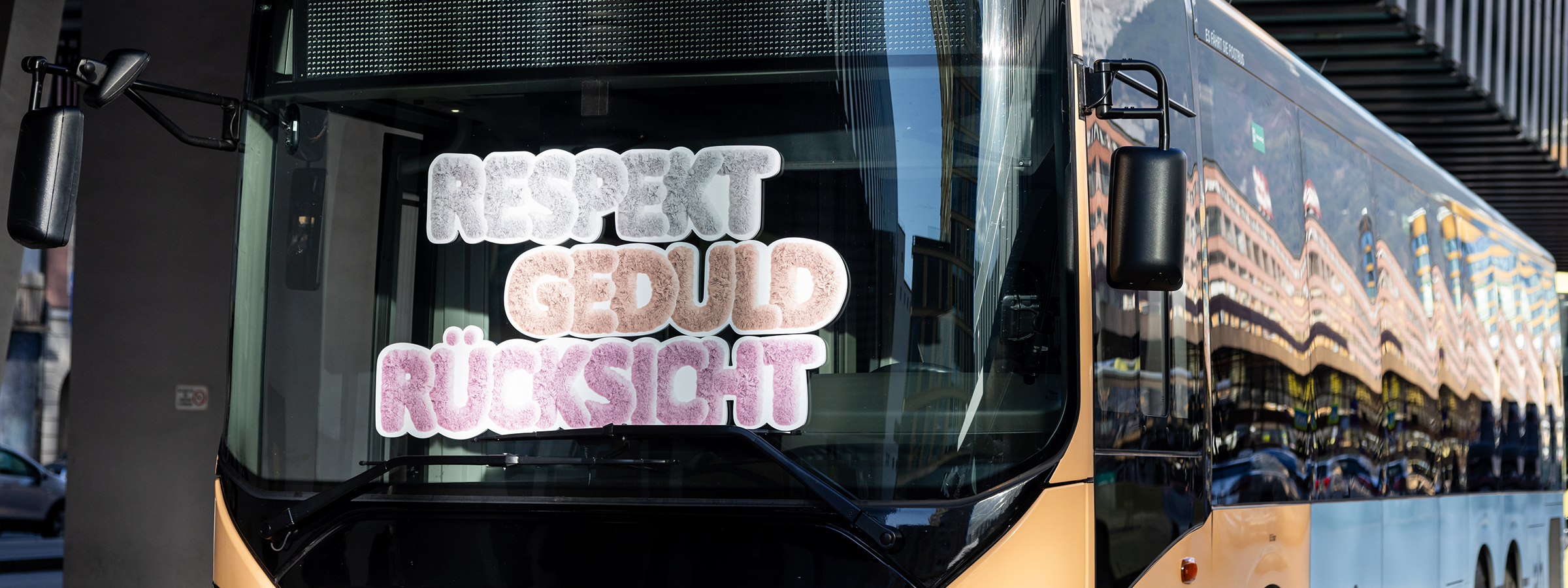Ein Nahverkehrsbus mit reflektierender Frontscheibe zeigt die Worte "Respekt", "Geduld" und "Rücksicht" in großen, flauschig gestalteten Buchstaben auf seiner Scheibe. Die Umgebung spiegelt sich in der glänzenden Oberfläche des Busses wider.