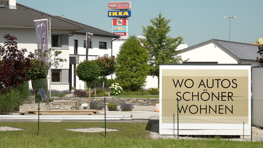 Das Foto zeigt ein Wohngebiet mit einem großen Schild. Auf dem Schild steht 'Wo Autos schöner wohnen'.