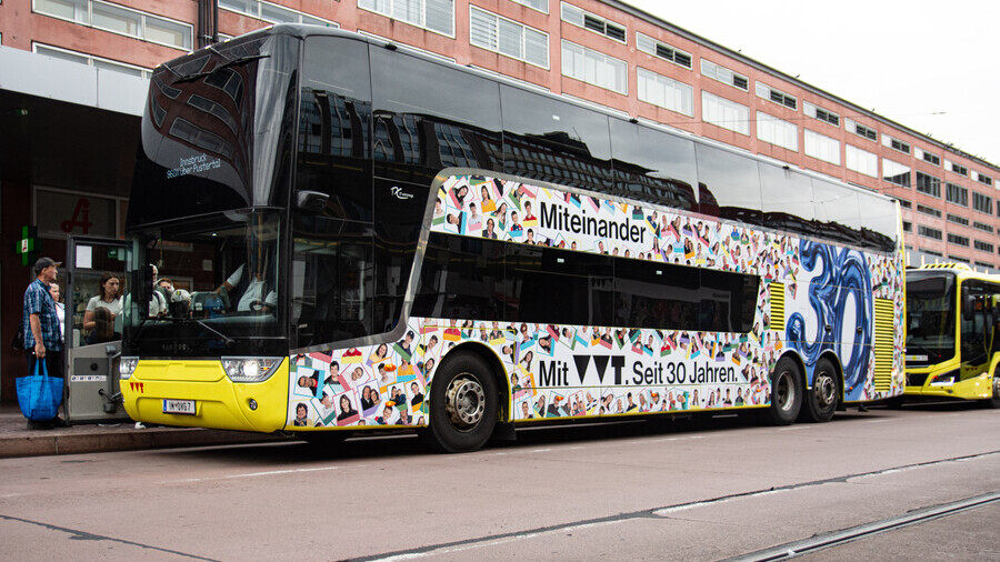 Man sieht den Doppelstockbus der Linie 960X von schräg vorne. Er ist mit unzähligen kleinen Porträtbildern beklebt.