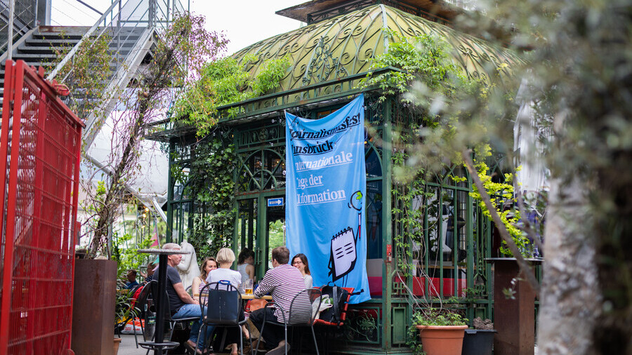 Eine Gruppe an Personen sitzt an einem Tisch vor einem grün bewachsenen Glashaus. Es handelt sich um das Treibhaus in Innsbruck. Ein Banner hängt vom Glashaus auf dem steht: "Journalismusfest Innsbruck. Internationale Tage der Information. 